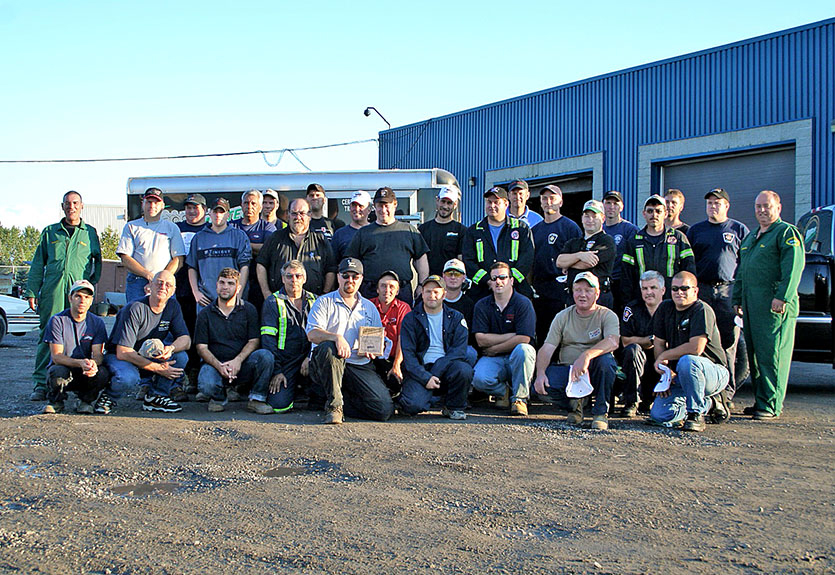 Group photo of  staff members standing in front of a blue industrial building and service vehicles