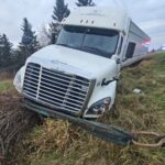 Front view of a white tractor-trailer leaning into a grassy roadside ditch.